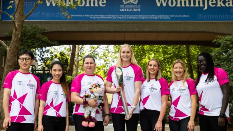 Seven baton bearers in magenta-and-white kit outside the University of Melbourne, one holding the Commonwealth Games baton